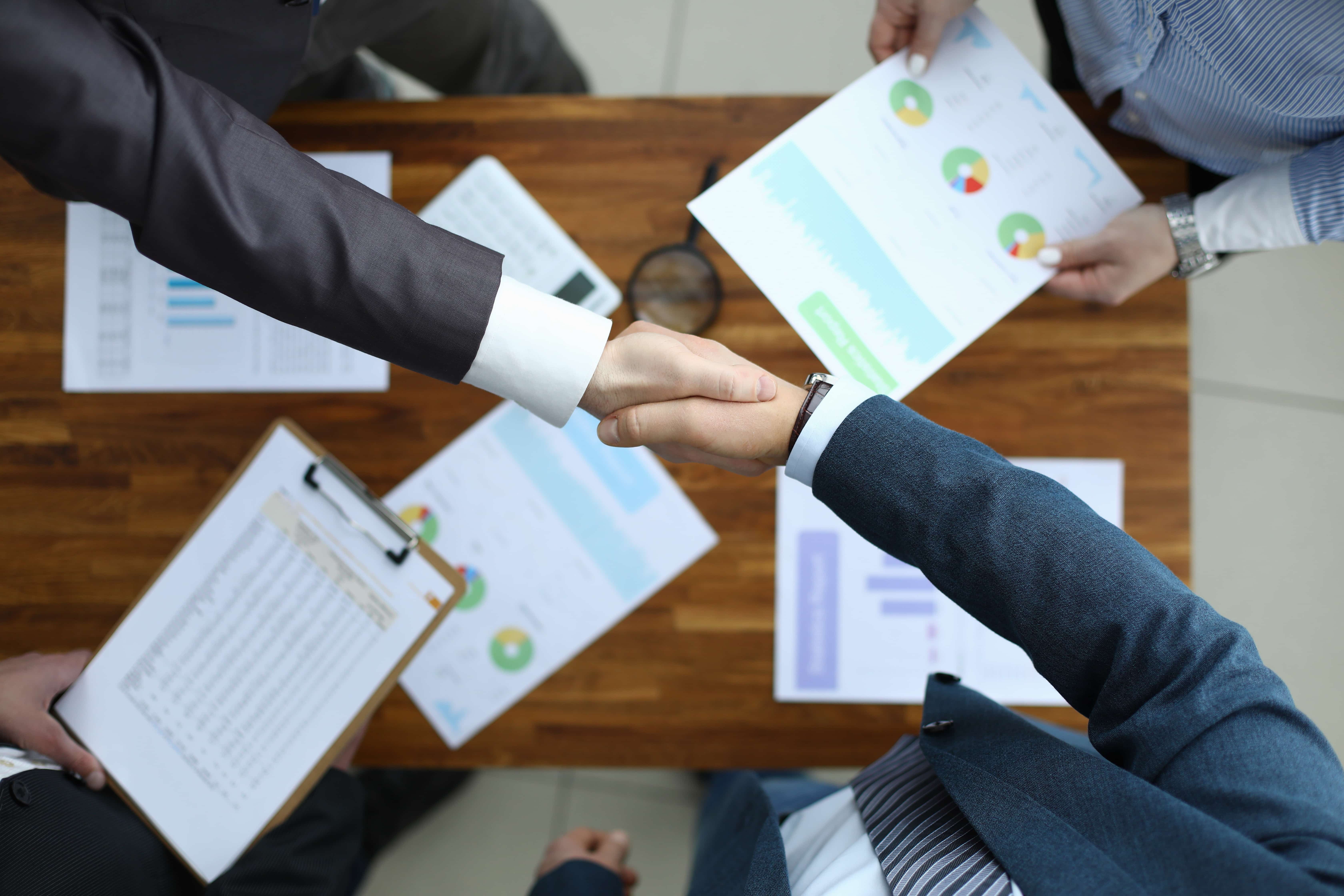 A handshake over a table with reports spread out. - Jerone Toledo Frente